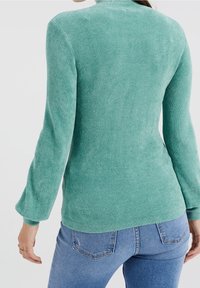 Grön ribbad stickad tröja med långa ärmar, med en hög krage och en mjuk textur. Bärs med blå jeans, vilket ger en avslappnad passform.