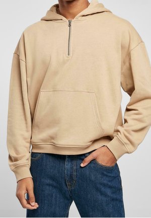 Beige hættetrøje med halvlukning og forlommer båret over blå denimjeans, med hænderne i lommerne, beskåret ved nakken og taljen.