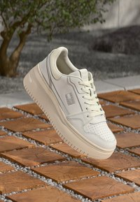 Baskets en cuir blanc avec semelle épaisse, coutures détaillées et lacets, flottant au-dessus d'un chemin pavé de carreaux en bois avec du gravier entre les dalles.