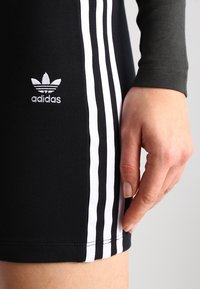 En hand vilar bredvid svarta Adidas-shorts med vita tre ränder och logotyp på sidan mot en plain bakgrund.