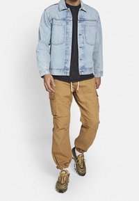 Lichtblauwe denimjack, tan kleurige cargobroek met elastische manchetten en gemusterde sneakers in groen en beige. Zwart t-shirt onder de jas.