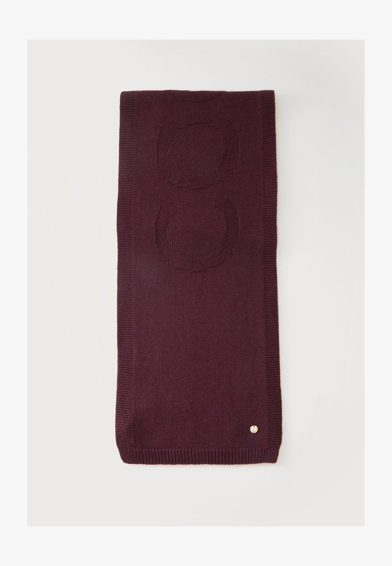 Écharpe tricotée de couleur violet foncé pliée, avec deux grands motifs circulaires tissés dans le tissu et un petit emblème métallique rond près du bord.