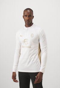 Weißes Fußballtrikot mit langen Ärmeln, goldenen Seitenpaneelen, goldener Logoplatzierung und Reißverschlusskragen. Hergestellt aus leichtem, strukturiertem Stoff mit sportlichem Design.