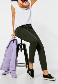 Persona con pantaloni verdi, top bianco senza maniche, sciarpa multicolore, sneakers nere, seduta su uno sgabello nero, mentre tiene una giacca viola chiaro.