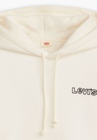 Krämfärgad huvtröja tillverkad av mjukt material, med en framficka, justerbara dragkedjor och en svart broderad "Levi's"-logga.