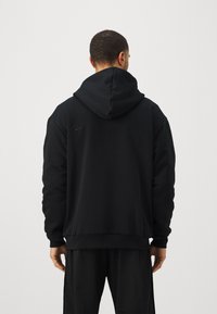 Melnā Nike kapuce ar plašu kapuci, ribotām piedurknēm un jostu. Iezīmēts ar mazām logotipa detaļām pie pleca. Mīksts auduma tekstūra.