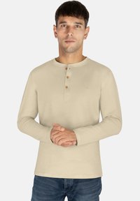 Beige Langarm-Henley-Shirt mit rundem Halsausschnitt, drei Holzknöpfen am Kragen und dezenten Stickdetails. Getragen mit blauen Jeans.