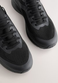 Black athletic shoes with a mesh upper, textured panels, and flat laces. Features a cushioned sole and rounded toe design for comfort.