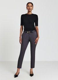 Schwarzer, kurzärmliger, gerippter Pullover kombiniert mit schmal geschnittenen grauen Hosen, akzentuiert durch einen schwarzen Gürtel mit goldener Schnalle und spitzen schwarzen Absätzen.