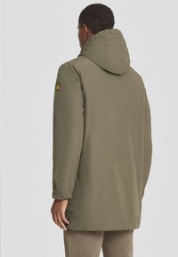 Abrigo acolchado verde oliva con capucha, presenta puños acanalados y un parche de logo visible en la manga. Textura suave y diseño de longitud media.