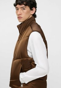 Braune gepolsterte Weste mit hohem Kragen, ausgestattet mit einer seitlichen Reißverschlusstasche und glatter, glänzender Textur. Tragen über einem weißen Langarmshirt.