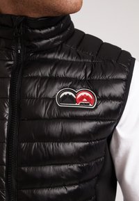 Gilet invernale nero con texture trapuntata, alto colletto e un'applique logo a contrasto con forme montane in rosso e nero. Chiusura con zip visibile.