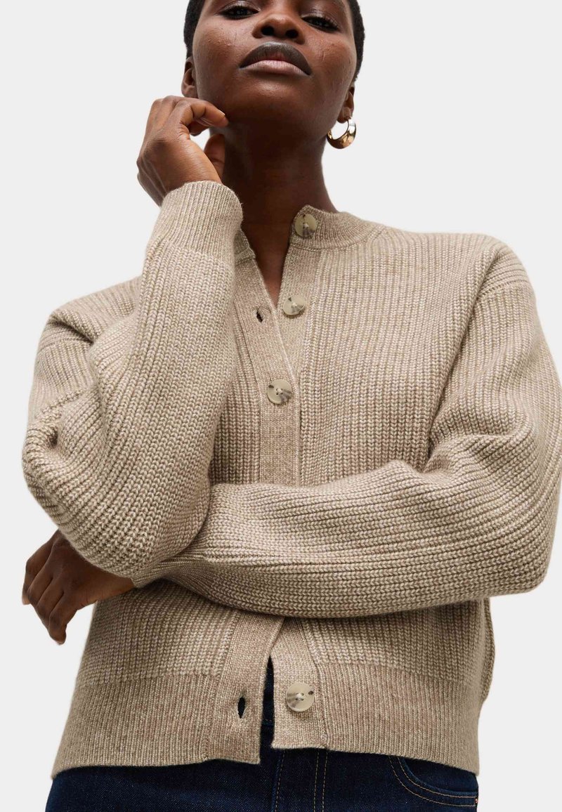 Cardigan en maille beige avec un motif texturé, doté d'un col montant, de boutons sur le devant et de poignets côtelés. Le matériau semble doux et chaud.