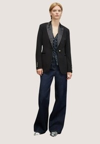 Blazer negro a medida con solapas de satén, un solo botón y detalles en tono dorado, combinado con una blusa estampada y pantalones de mezclilla oscuros de pierna ancha.