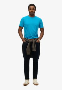 Lys blå kortærmet T-shirt, mørke jeans, grå sweatshirt bundet om taljen, beige sneakers og et armbåndsur på venstre håndled.