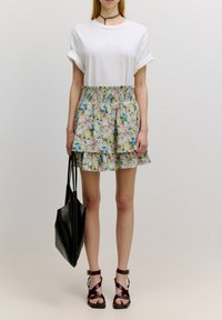 Wit kortoef t-shirt gecombineerd met een mini-jurk in pasteltinten met bloemenprint en meerdere lagen. Een zwarte schoudertas en bruine sandalen met bandjes maken de look compleet.