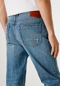 Heledad sinine teksarihm, mis on sirge jalgade lõikega, viie taskuga ja pruun nahkplekk vöökoosal, millel on logo detail.