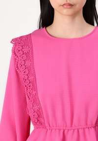 Robe rose en tissu texturé, avec des détails en dentelle sur une épaule et une taille froncée. Encolure ronde et manches longues.