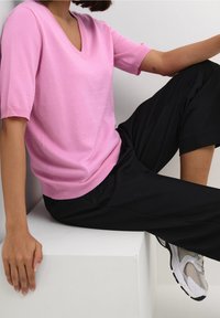Persona che indossa una maglietta rosa a maniche corte con scollo a V, pantaloni neri e sneaker bianche, seduta su un blocco bianco contro un muro bianco.