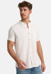 Jonge man met krullend haar, gekleed in een wit overhemd met korte mouwen en blauwe jeans, die iets opzij kijkt.