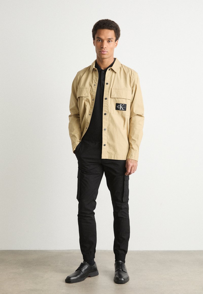 Camicia beige chiaro con due tasche frontali e logo nero applicato, abbinata a pantaloni cargo neri e scarpe nere. Design semplice e casual.