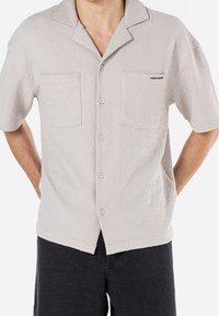 Koszulka z krótkim rękawem w jasnoszarym kolorze, z teksturowanym wzorem, z kołnierzykiem, dwiema kieszeniami na piersi i guzikami z przodu. Ozdobiona logo "PEGADOR".