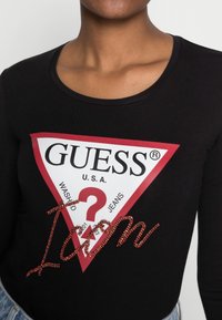 Czarna koszula z długim rękawem z białym trójkątnym logo z napisem "GUESS" i "U.S.A.", ozdobiona czerwonymi rhinestonami układającymi się w słowo "Icon."