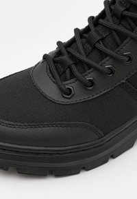 Black athletic shoe with a textured fabric upper, smooth rubber accents, and a thick sole. Features black laces and reinforced eyelets.