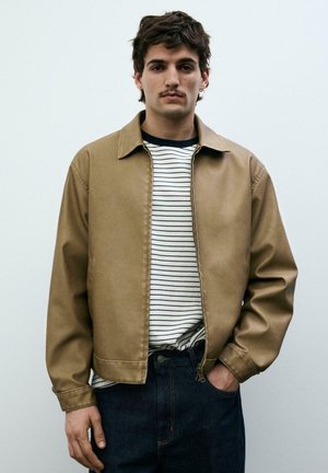 Bărbat tânăr cu păr scurt și închis la culoare și mustață, purtând o jachetă bej peste o cămașă albă cu dungi negre și blugi închiși la culoare, stând cu o mână în buzunar.