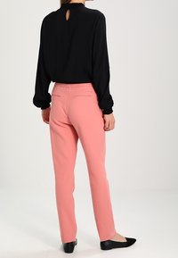 Zwarte blouse met lange mouwen en een ronde halslijn met een sleutelgatopening aan de achterkant. Roze, op maat gemaakte broek met achterzakken en slim fit. Zwarte flats.
