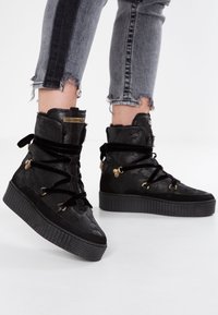 Mustat korkeat tennarit, joissa on teksturoitu päällinen, nauhakiinnitys, samettiyksityiskohtia, kultaiset metalliosat ja paksu kumipohja.