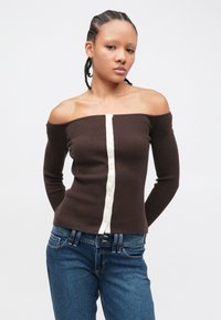 Haut marron côtelé, épaules dénudées, manches longues, fermeture boutonnée à l'avant avec des boutons crème. Associé à un jean denim bleu.