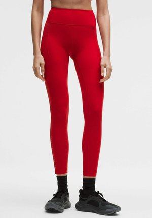 Leggings rouge taille haute slim avec poches latérales portés par une personne debout, assortis à des chaussettes noires et des chaussures de sport noires.