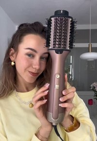 Donna con un maglione giallo che tiene in mano un grande pettine per capelli color rosé con pulsanti, sorridendo in una stanza ben illuminata con piante decorative.