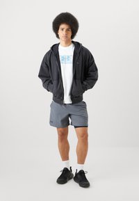 Čierna mikina na zips cez biele tričko s svetlomodrým logom, skombinovaná so šedými šortkami a čiernymi športovými topánkami. Viditeľné biele ponožky.