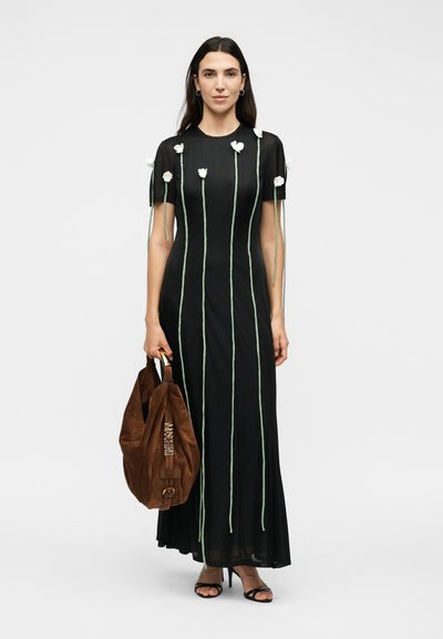 Γυναίκα που φοράει μαύρο μακρύ φόρεμα με κάθετα πράσινα στελέχη και λευκά λουλούδια, κρατώντας μια μεγάλη καφέ σουέντ τσάντα, στέκεται σε λευκό φόντο.
