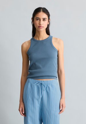 Młoda kobieta z długimi ciemnymi włosami, ubrana w niebieski bezrękawnik i jasnoniebieskie, paskowane spodnie ze ściągaczem, stojąca na jednolitym szarym tle.