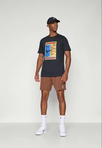 Muž oblečený v černé kšiltovce Nike, černém tričku s potiskem, hnědých šortkách, bílých ponožkách Nike a bílých teniskách stojí před jednoduchým pozadím.