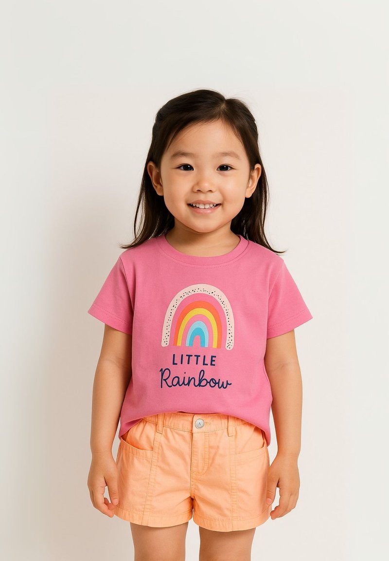 Rosa Baumwoll-T-Shirt mit dem Aufdruck "Little Rainbow", das ein mehrfarbiges Regenbogendesign zeigt, kombiniert mit orangefarbenen Shorts. Kind lächelt.