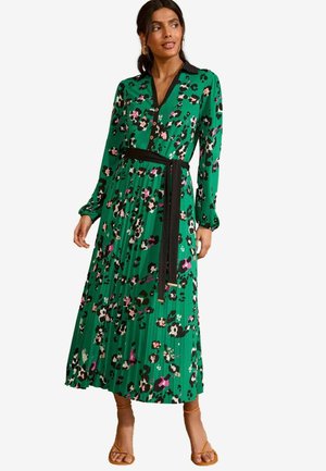 Maxi abito floreale verde con accenti neri, chiusura a bottoni frontale, maniche lunghe e una cintura nera legata in vita; texture plissettata su tutta la superficie.