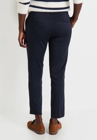 Pantalons tailleur bleu marine avec une coupe ajustée, dotés de deux poches arrière, d'un ourlet à revers et d'une texture lisse ; associés à des mocassins en daim marron.