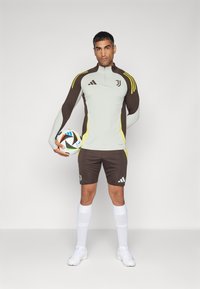 Haut de sport à manches longues gris avec des accents marron et jaune, associé à un short marron. Le joueur tient un ballon de soccer multicolore. Chaussettes et chaussures blanches.