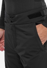 Schwarze Outdoor-Hosen aus wasserabweisendem Material, mit einem Reißverschluss, zwei Knopfverschlüssen und einem Klettverschlussverstellriemen an der Taille.
