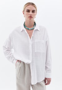Camisa blanca de botonadura en tela texturizada con mangas largas y bolsillo en el pecho, combinada con pantalones beige claro y collar de cuentas azules.
