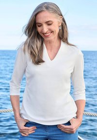 Witte, lichte V-hals top met driekwart mouwen, gecombineerd met blauwe jeans, buiten bij water. Model heeft lang grijs haar.