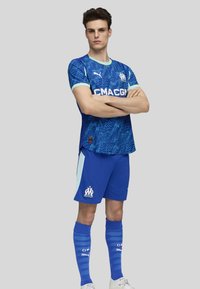 Zila futbola krekls ar strukturētu, teksturētu dizainu, kurā ir gaiši zili akcenti un balti sponsoru/logo detalizējumi. Pāris kopā ar ziliem šortiem un zeķēm.