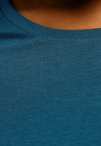 Primo piano della parte superiore del torace e del collo di una persona che indossa una maglietta a girocollo a coste color turchese.