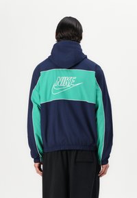 Športna jakna v mornarsko modri in teal barvi z kapuco in elastičnimi manšetami. Ima izstopajoč bel Nike logotip na hrbtni strani in sproščen kroj.