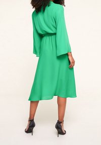 Vrouw staat met haar rug naar ons gekeerd, gekleed in een felgroene midi-jurk met lange mouwen en zwarte hoge hakken met enkelbandjes.