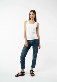 Ein weißes ärmelloses Tank-Top, Jeans aus Denim und schwarze Sandalen. Das Tank-Top hat einen runden Halsausschnitt, die Jeans haben eine lockere Passform und die Sandalen sind mit verstellbaren Riemen ausgestattet.